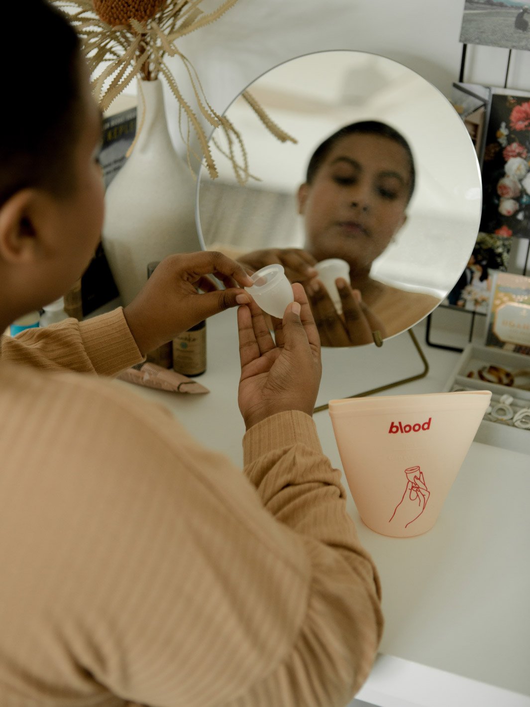 woman sitting in front of a vanity mirror looking at a menstrual cup. sanitising pouch is on the table next to her.